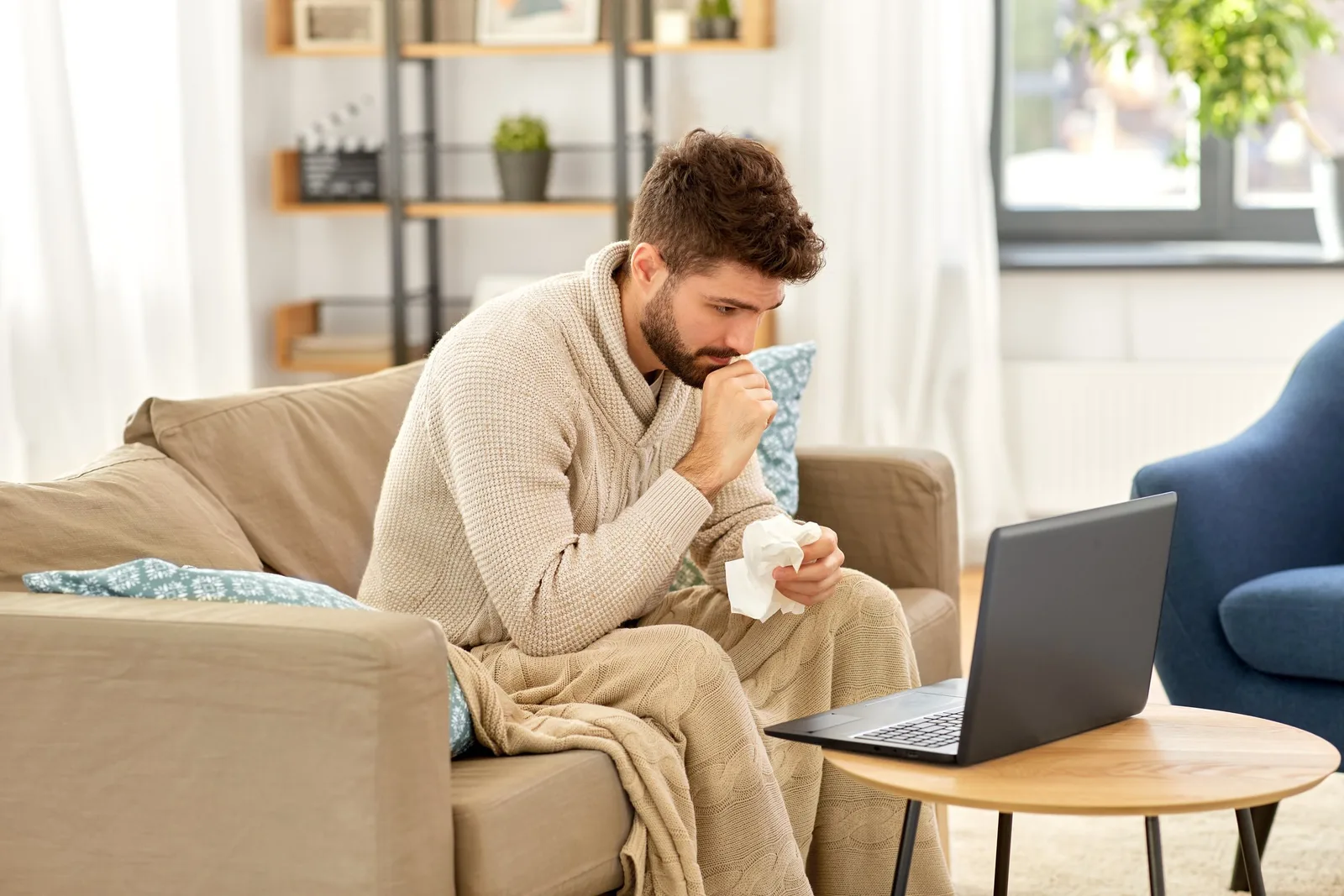 Person med förkylning hemma i soffan med te och dator, illustrerar sjukpenning och karensavdrag 2026